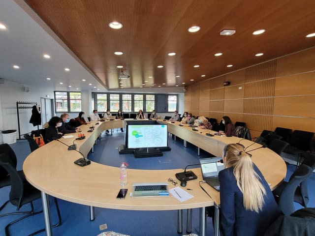 Photo d'une grande salle claire, avec une table ovale et une quinzaine de personnes assises autour