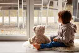 Photographie qui met en scène un enfant qui regarde par la fenêtre et qui tient son doudou.
