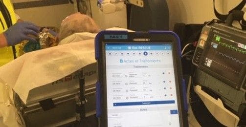L'écran de l'application s'affiche sur une tablette avec en arrière plan une main gantée et la tête d'un patient alité